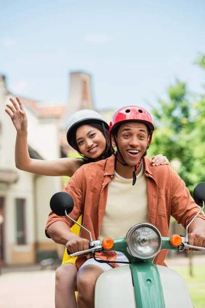 快乐的亚洲女人一边和兴奋的非洲男人一起骑摩托车一边挥手 — 图库照片