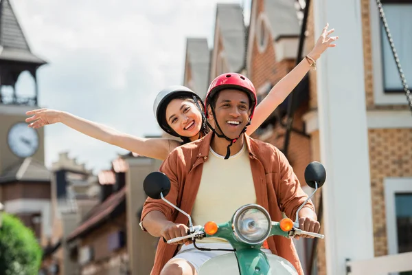 cheerful asian woman raising hands while riding scooter with african american man