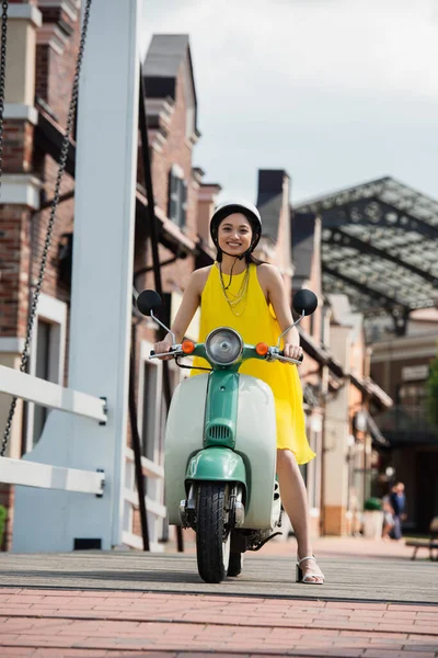 幸せなアジアの女性で黄色のサンドレスに乗るスクーターで都市の通り — ストック写真