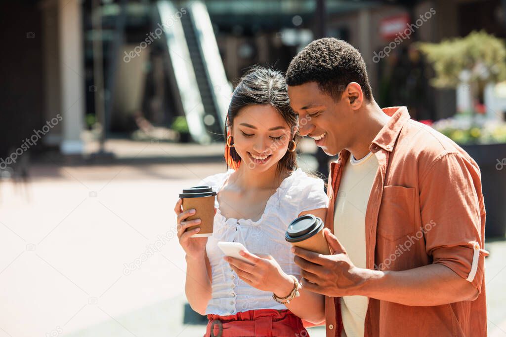 cheerful interracial couple with paper cups looking at mobile phone outdoors