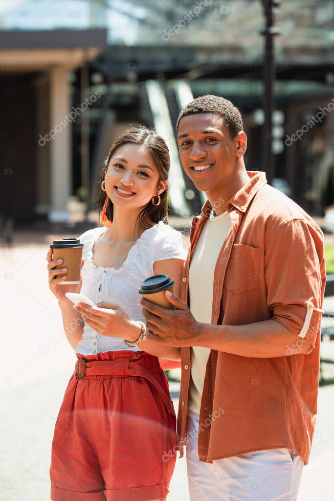cheerful interracial couple with coffee to go smiling at camera outdoors
