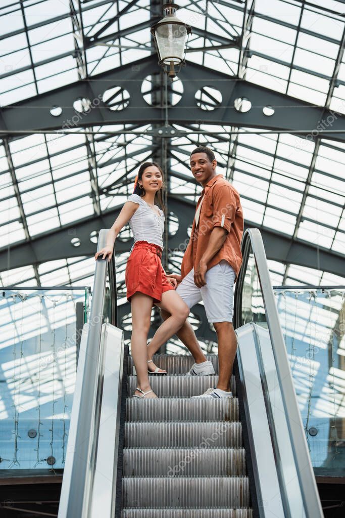 low angle view of interracial couple in stylish summer clothes smiling at camera on escalator