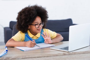 Afro-Amerikalı kız dizüstü bilgisayara el sallıyor ve evdeki deftere yazıyor. 