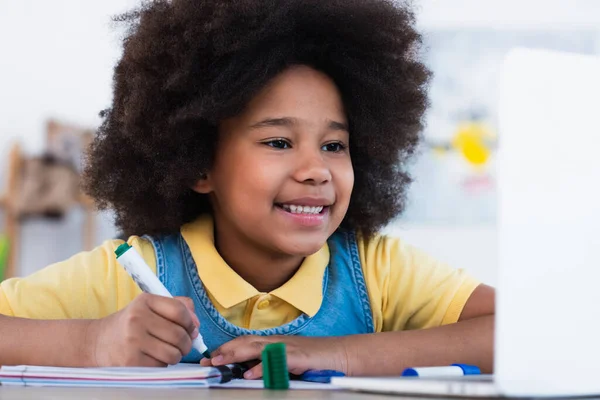 Sonriente Niño Afroamericano Escribiendo Cuaderno Cerca Computadora Portátil Borrosa Casa — Foto de Stock