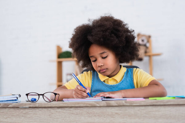 African american girl writing on notebook near eyeglasses on table 