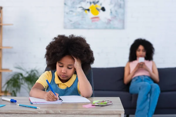 Niño Afroamericano Escribiendo Cuaderno Cerca Madre Borrosa Con Teléfono Inteligente — Foto de Stock