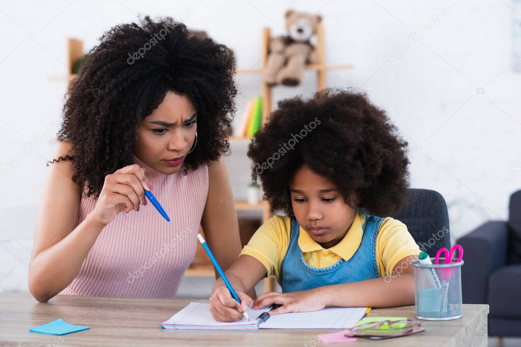 Angry african american mother pointing with pen near daughter writing on notebook at home 