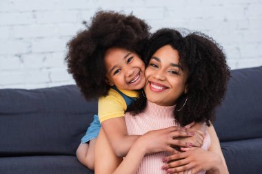 Neşeli Afro-Amerikalı çocuk evde kanepede gülümseyen anneyi kucaklıyor. 