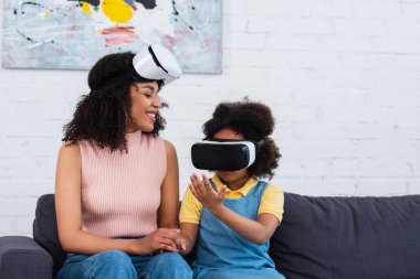 Gülümseyen Afrikalı Amerikalı anne video oyunu oynayan kızına VR kulaklıkla bakıyor. 