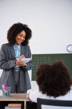 Genç Afro-Amerikan öğretmeni bulanık okul çocuğu ve kırtasiyesinin yakınındaki deftere yazı yazıyor. 