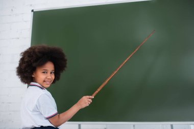 Gülümseyen Afrikalı Amerikalı okul çocuğu kameraya bakıyor ve sınıftaki tahtayı işaret ediyor. 