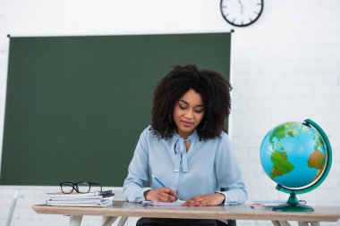 Sınıfta dünyanın yakınındaki not defterine yazı yazan Afrikalı Amerikalı öğretmen. 