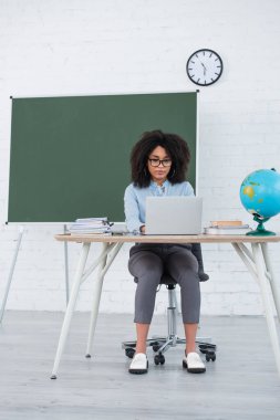 Gözlüklü Afrikalı Amerikalı öğretmen okulda kitapların yanında dizüstü bilgisayar ve dünya kullanıyor. 