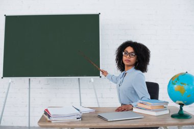 Kara tahtayı işaret eden ve kitapların, laptopun ve dünyanın yakınındaki kameraya bakan Afrikalı Amerikalı öğretmen. 