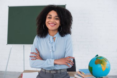 Sınıfta kollarını kavuşturup kameraya gülümseyen Afrikalı Amerikalı öğretmen. 