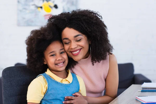 Madre Afroamericana Sonriendo Mientras Abraza Hija Cerca Cuadernos Mesa — Foto de Stock