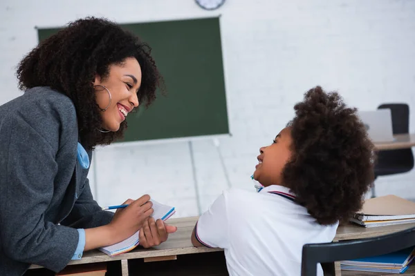 Sonriente Profesor Afroamericano Escribiendo Cuaderno Cerca Alumno Aula — Foto de Stock