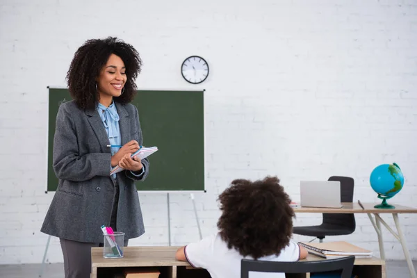 Profesora Afroamericana Positiva Escribiendo Cuaderno Cerca Escolar Borroso Durante Lección —  Fotos de Stock