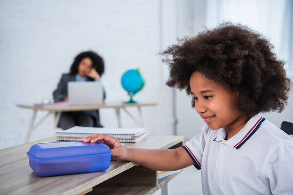 Smiling african american kid holding lunch box near blurred notebooks in classroom 
