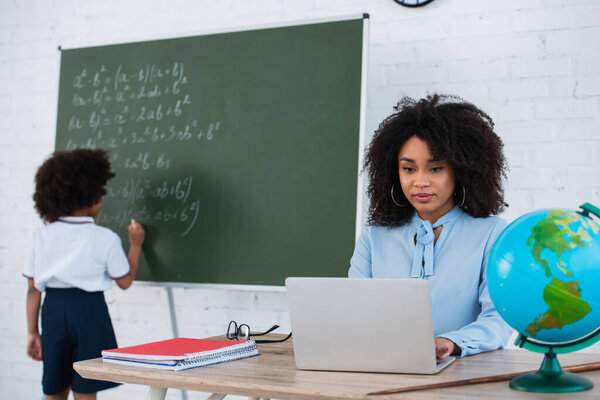 African american teacher using laptop while pupil writing on chalkboard on blurred background 
