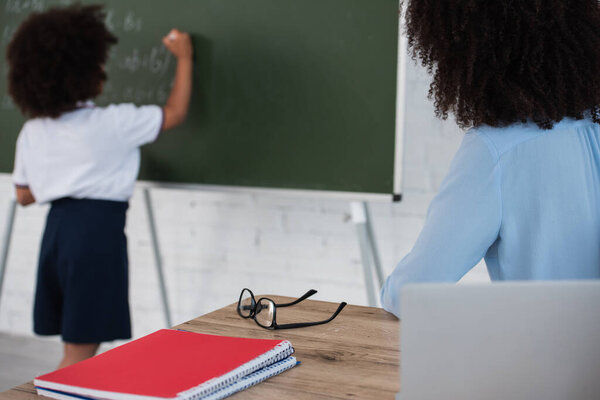 African american teacher sitting near laptop and blurred pupil writing on chalkboard 