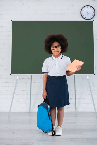 Smiling african american girl holding book and backpack in classroom 