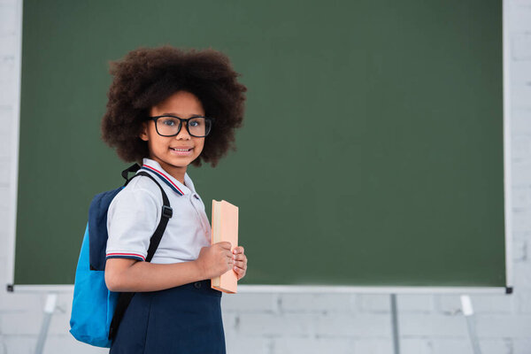 Happy african american child with backpack and book standing near chalkboard in school 