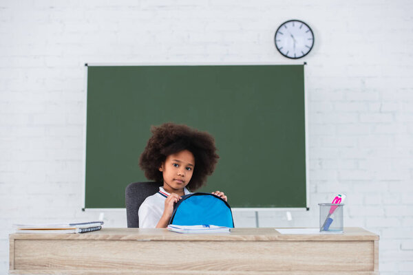 African american schoolchild with backpack looking at camera near desk in classroom 