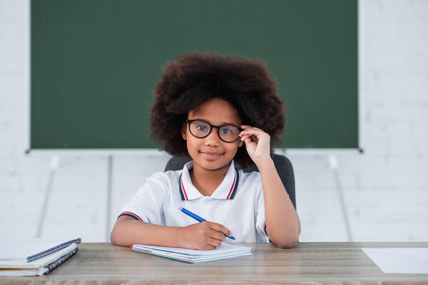 Smiling african american child holding eyeglasses and pen near notebooks on desk in school 