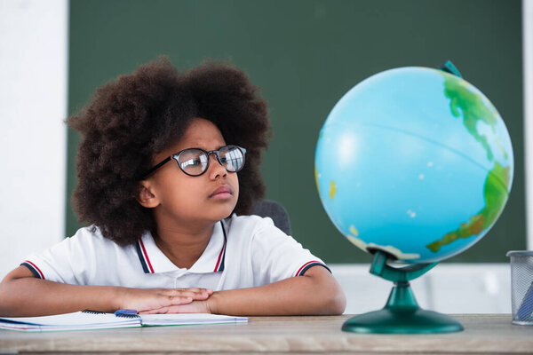 African american schoolchild in eyeglasses looking at blurred globe 