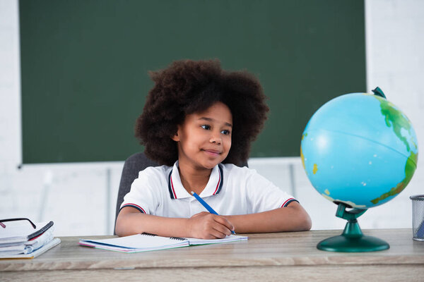 Smiling african american pupil writing on notebook near blurred globe on desk 