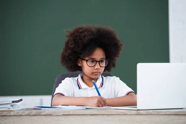 Niño Afroamericano Escribiendo Cuaderno Cerca Una Computadora Portátil Borrosa Aula — Foto de Stock