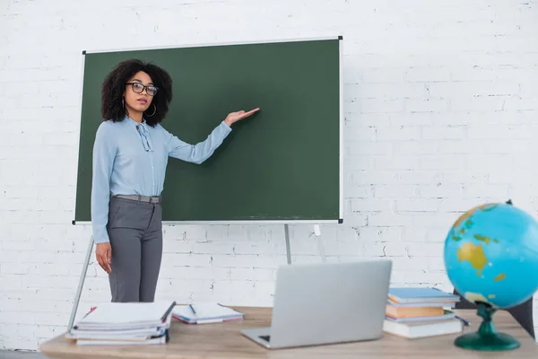 African american teacher pointing at chalkboard near laptop during video chat in school