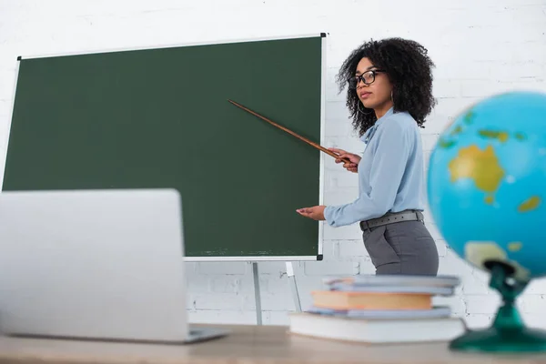 Profesor Afroamericano Apuntando Pizarra Cerca Computadora Portátil Borrosa Durante Lección —  Fotos de Stock