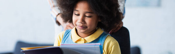 African american kid looking at notebook near blurred mother, banner 