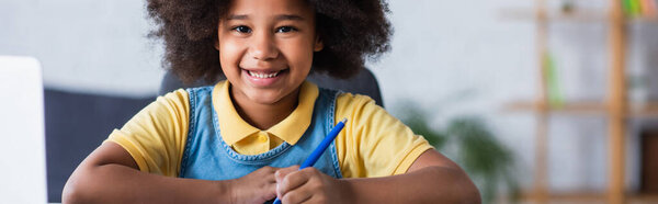 Happy african american kid holding pen near blurred laptop, banner 