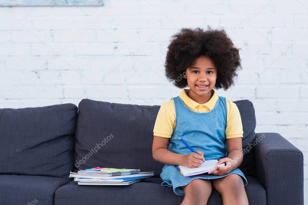 African american girl smiling at camera while doing schoolwork on couch 