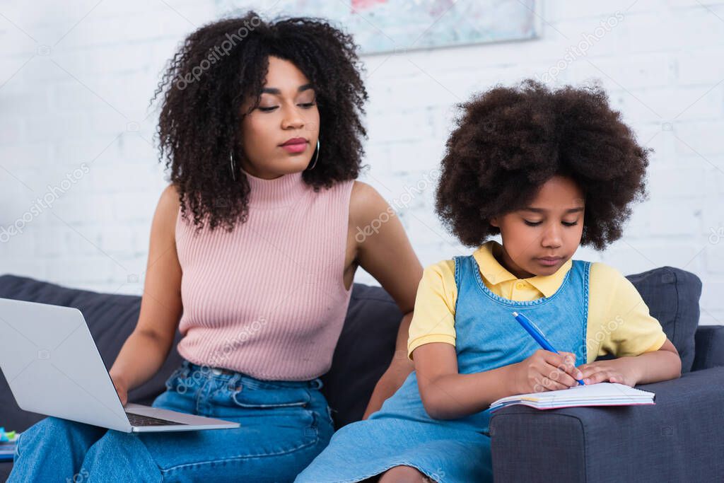 African american kid writing on notebook near blurred mother with laptop on couch 