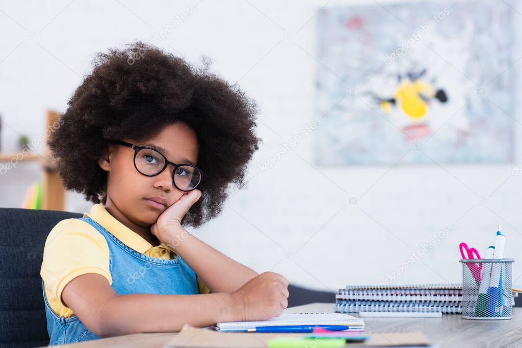 African american girl in eyeglasses looking at camera near copy books and stationery at home 