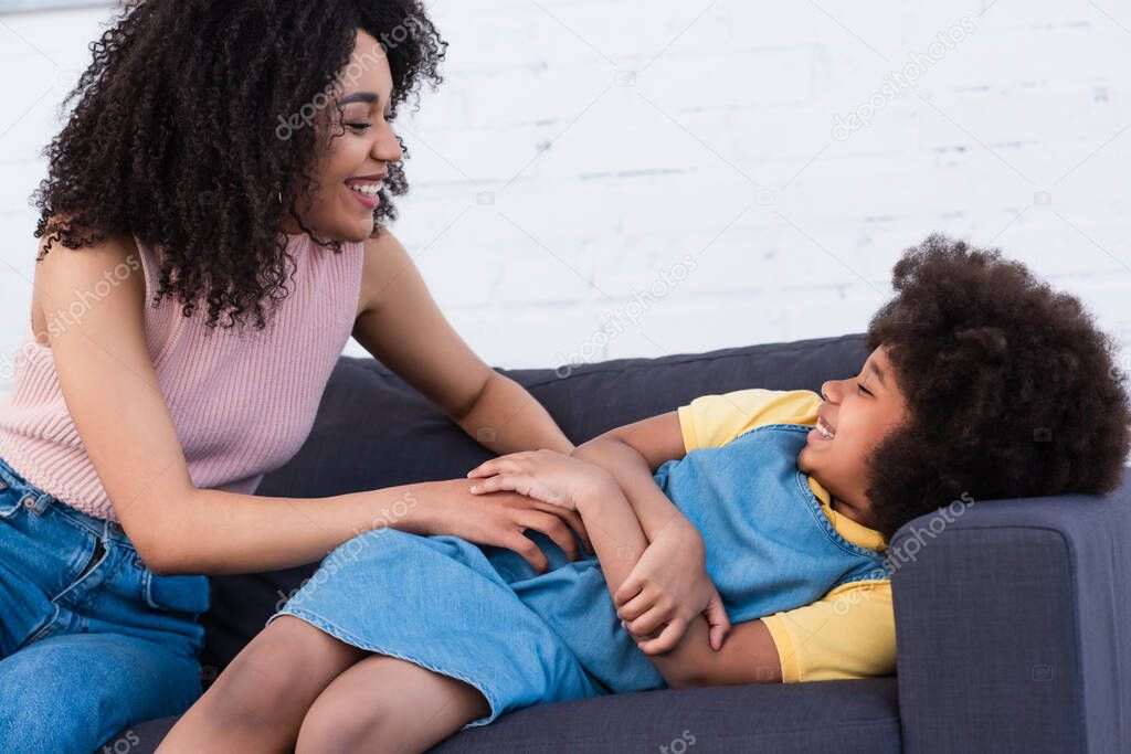 Cheerful african american woman tickling daughter on couch 