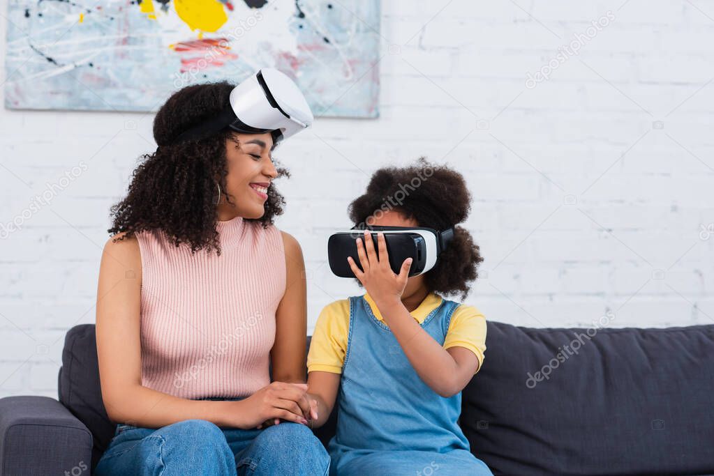 Smiling african american parent looking at daughter in vr headset at home 