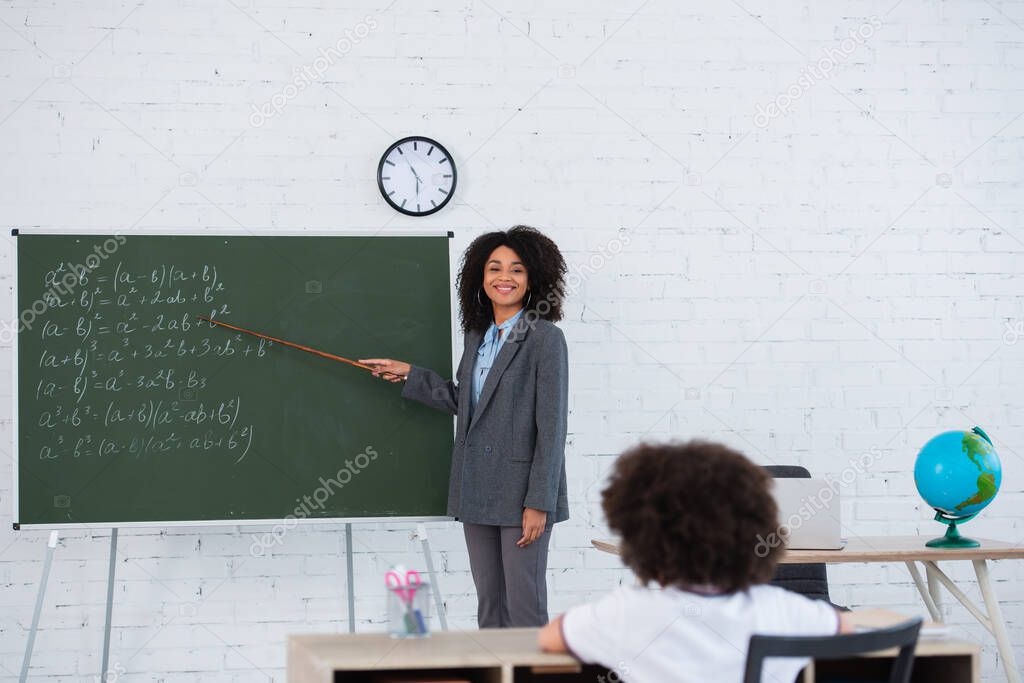 Smiling african american teacher with pointer standing near chalkboard and blurred schoolkid in classroom 