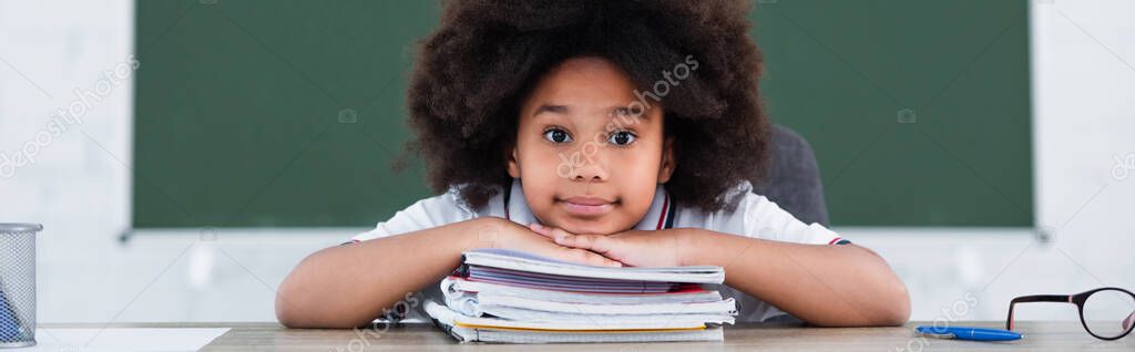 African american schoolkid looking at camera near notebooks on desk in classroom, banner 