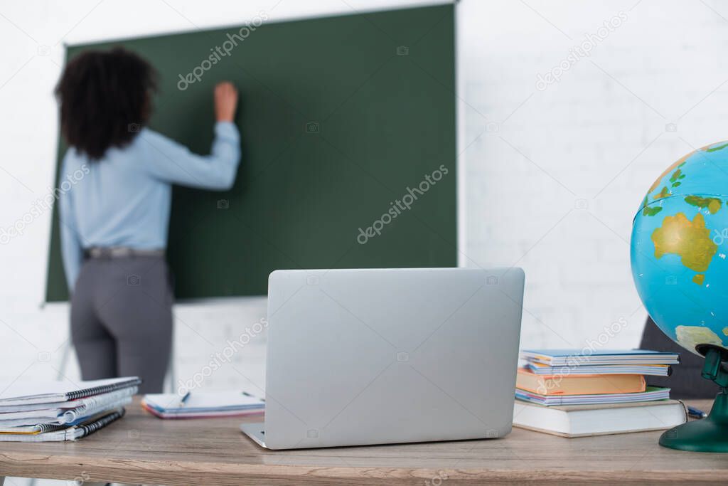 Laptop, globe and notebooks on table near blurred african american teacher writing on chalkboard 