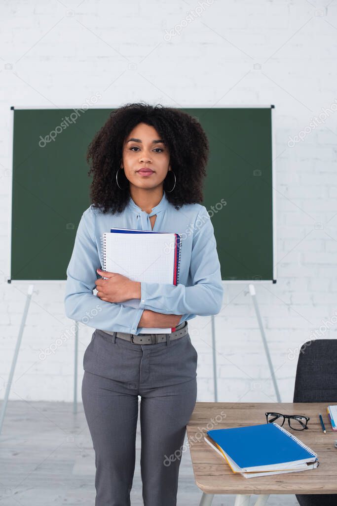 African american teacher holding notebooks and looking at camera in classroom 