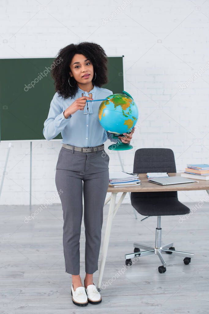 African american teacher pointing at globe and looking at camera in school 