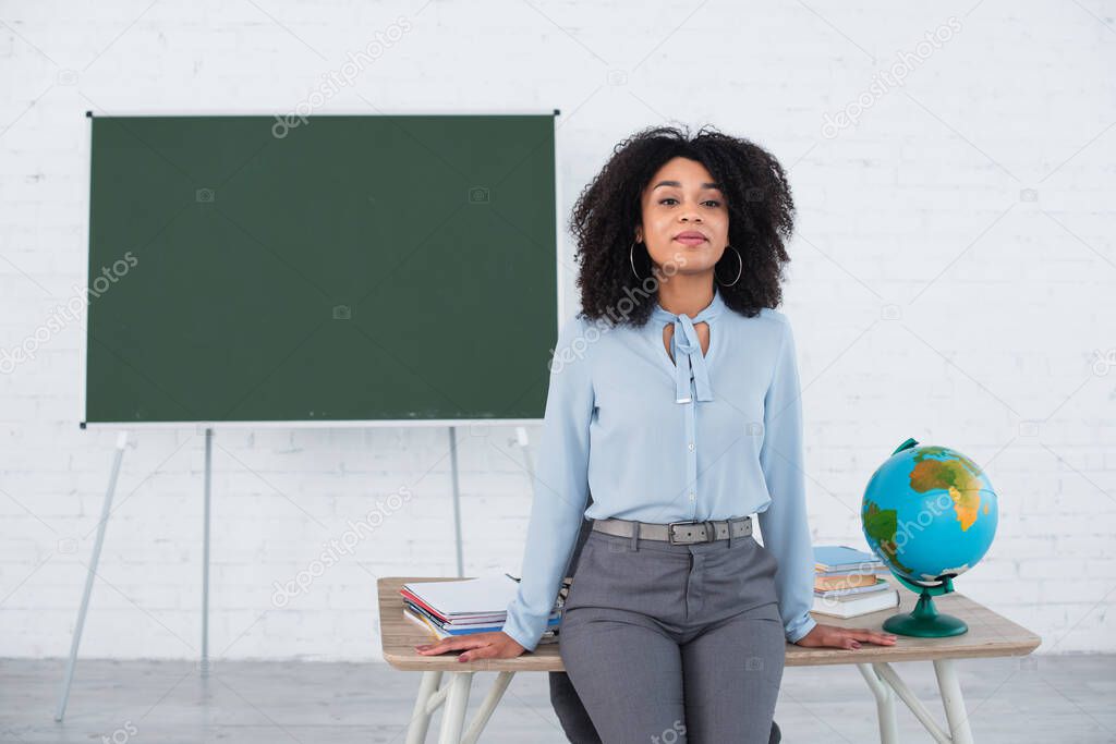 Young african american teacher standing near globe and notebooks on working table 