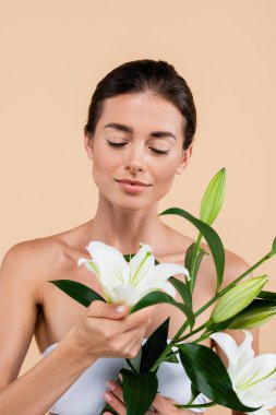 charming woman with naked shoulders looking at white lily flowers isolated on beige, beauty concept clipart