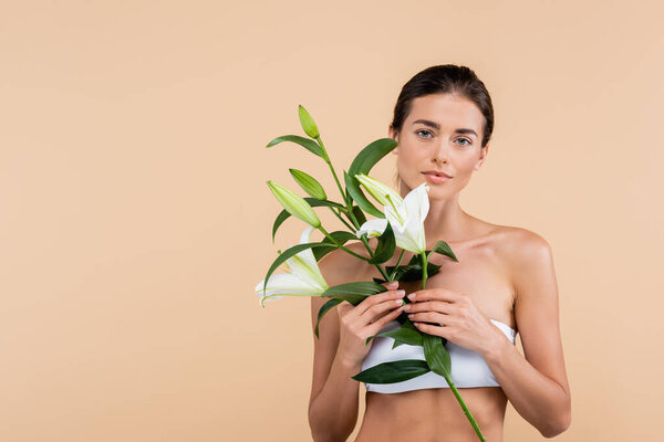 charming woman with white lily flowers looking at camera isolated on beige, beauty concept
