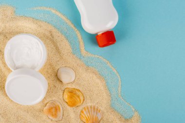 Kum ve mavi yüzey üzerinde deniz kabuklarının yanında güneş kremi ve kozmetik kremin üst görüntüsü